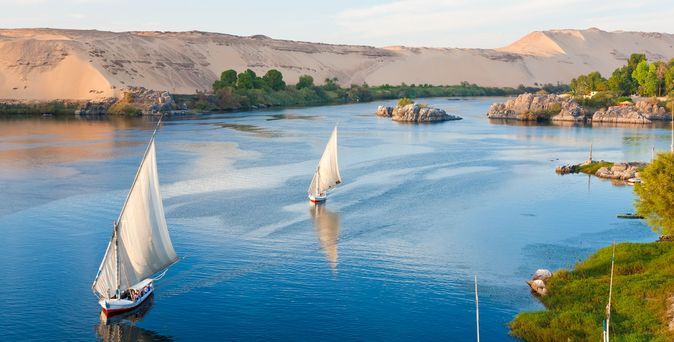 Traditionelle Feluccas segeln sanft über den Nil, umgeben von grünen Ufern und sandigen Dünen – ein Bild zeitloser Ruhe.