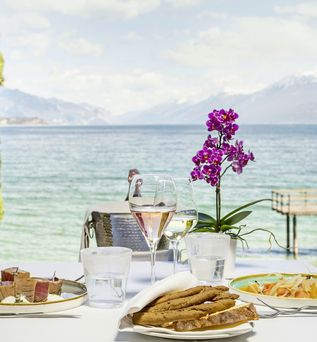 Tischgedeck mit Speisen, Wein und Orchidee auf einer Restaurantterrasse mit Seeblick und Anlegesteg im Hintergrund.