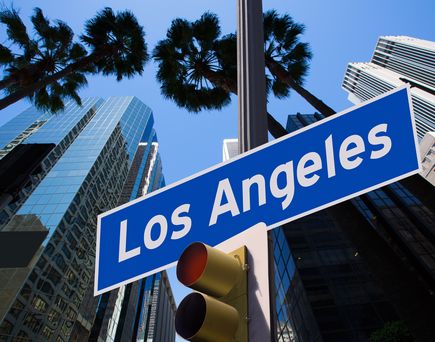 Am unteren Rand ist das rote Licht einer Ampel zu erkennen. Darüber ein blaues Schild mit weißer Schrift 'Los Angeles'. Umgeben von Hochhäusern und hohen Bäumen. Blauer Himmel.