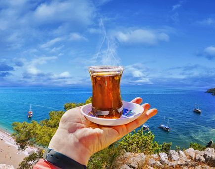 Ein Gläs türkischer Tee in der Hand, oberhalb der türkisblauen Bucht, umgeben von etwas Wald und Bergen. Blauer Himmel. Alles aus einer Panromasicht.