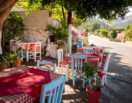 Eine Taverne am Wegesrand bzw. einer Orts-Ende-Straße mit vielen Bäumen. Weiße Stühle und ein mit rot-karierter Tischdecke bedeckter Tisch. Sehr einladend.