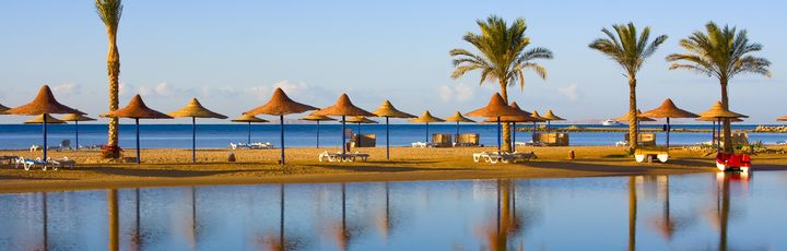 Ein Sandstrand mit Reihen von Strohsonnenschirmen und Liegestühlen, die sich an einem sonnigen Tag an einem Hotelresort in Hurghada, Ägypten, widerspiegeln.