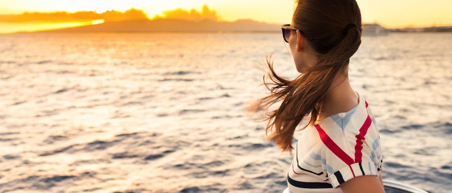 Eine Frau steht an der Reling eines Schiffes und blickt auf den Sonnenuntergang über dem Meer. Ihre Haare wehen im Wind.