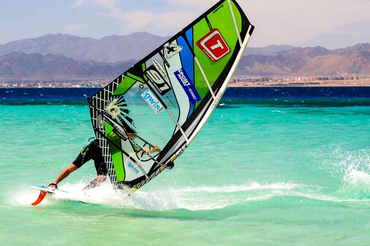 Ein Windsurfer auf dem türkisfarbenen Wasser, der ein grünes Segel mit einer roten und weißen Grafik auf einem sonnigen Tag benutzt.