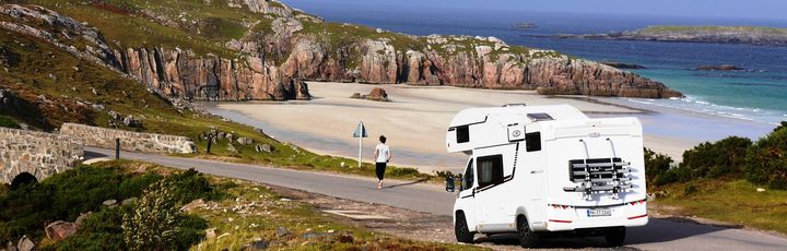 Ein Camper steht an einer Straße nähe einem Strand mit Felsen und ein Mann läuft in Richtung Strand weg vom Camper