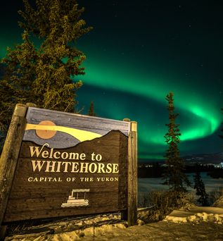 Das Willkommensschild für Whitehorse, Yukon, Kanada, steht prominent in einer grünen Landschaft mit Bäumen und einem weiten, blauen Himmel, der Reisende begrüßt.