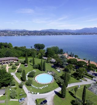 Ferienanlage mit gepflegtem Garten, rundem Pool und Zypressen direkt am Ufer eines großen Sees mit Blick auf die Berge.
