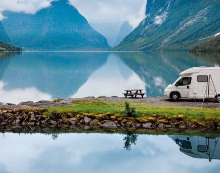 Weißer Campervan steht am Ufer eines ruhigen Sees, umgeben von grünen Bergen, spiegelt sich im Wasser.