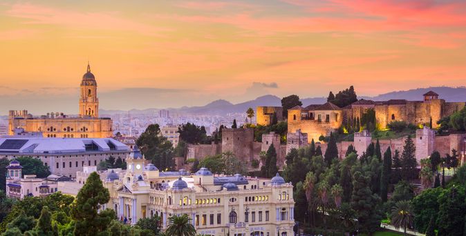 Blick auf die Altstadt von Malaga. Zu sehen sind wunderschöne, teils alte Bauten, umgeben von viel Grün. Kathedrale im Hintergrund bei Sonnenuntergang