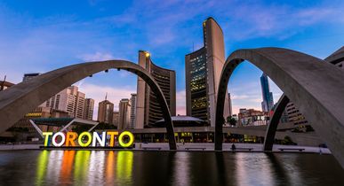 Blick auf die New City Hall, das neue Rathaus, mit seinen zwei geschwungenen Türmen. Im Vordergrund der beleuchtete, ikonische TORONTO Schriftzug bei Sonnenaufgang.