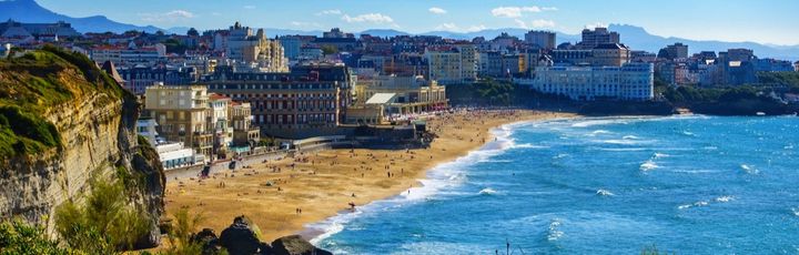 Der breite Sandstrand von Biarritz, Frankreich, mit Wellen, die auf das Ufer treffen, und der Stadt im Hintergrund unter einem bewölkten Himmel.