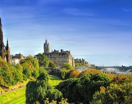 Eine beeindruckende Panoramaansicht der Princes Street Gardens in Edinburgh, Schottland, mit dem majestätischen Edinburgh Castle, das auf einem Hügel thront, und dem Scott Monument im Vordergrund, unter einem klaren blauen Himmel.