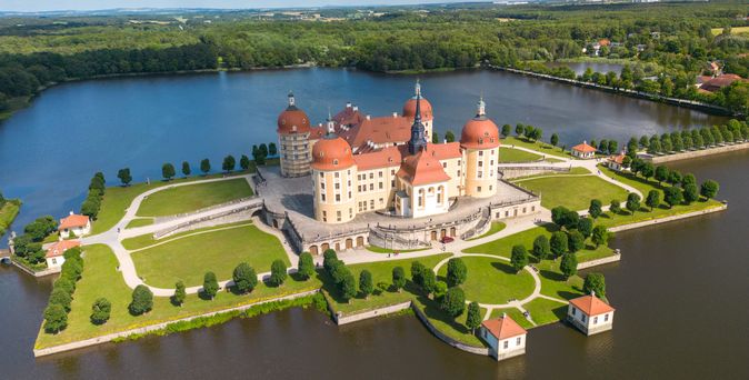 Ein Luftbild des beeindruckenden Moritzburg Schlosses, das malerisch auf einer Insel in einem See liegt und von gepflegten Gärten und einem weitläufigen Wald umgeben ist.
