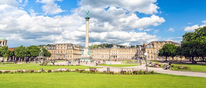 Blick auf den Schlossplatz in Stuttgart mit Jubiläumssäule, gepflegtem Rasen und dem Neuen Schloss unter blauem Himmel mit weißen Wolken.