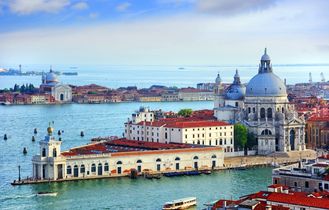 Die imposante Basilika Santa Maria della Salute in Venedig, Italien, mit ihrer markanten Kuppel und den venezianischen Gebäuden am Canal Grande, unter einem blauen Himmel.   Sources