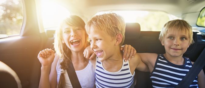 Drei fröhlich lachende Kinder, angeschnallt auf dem Rücksitz eines Autos, bei sonnigem Wetter.