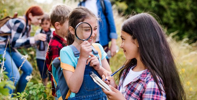 Zwei Kinder spielen in der Natur, ein Mädchen zeigt dem Jungen etwas auf einer Baumwurzel.