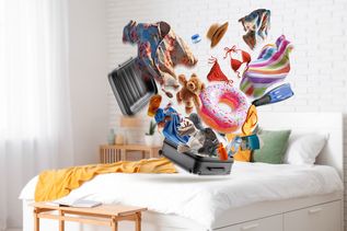 Motiv Reiserücktrittskostenversicherung ADAC Bett auf dem ein Koffer Steht aus dem Kleidungsstücke fliegen