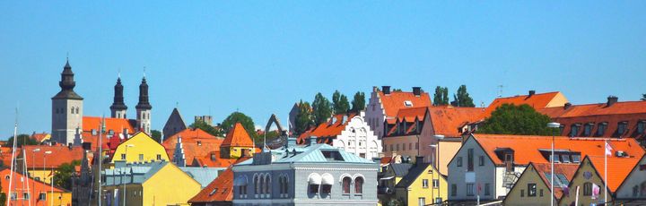 Blick auf die bunten Häuser von Visby, ein Steg am Wasser, Cafe und Sitzmöglichkeiten dort. Blauer Himmel.