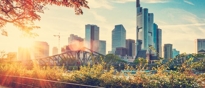 Sonnenuntergang über der Skyline von Frankfurt am Main mit modernen Hochhäusern, dem Mainufer und herbstlich gefärbtem Grün im Vordergrund.