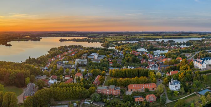 Eine malerische Luftaufnahme von Plön in Schleswig-Holstein, die das Plöner Schloss auf einer Insel im Großen Plöner See zeigt, umgeben von einer grünen Landschaft und dem Plöner See, unter einem strahlend blauen Himmel.