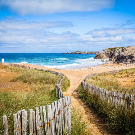 Eine erhöhte Ansicht eines weitläufigen Sandstrandes mit sanften Dünen im Vordergrund, die von einem hölzernen Zaun begrenzt werden. Das türkisfarbene Meer erstreckt sich bis zum Horizont unter einem hellblauen Himmel mit vereinzelten Wolken.