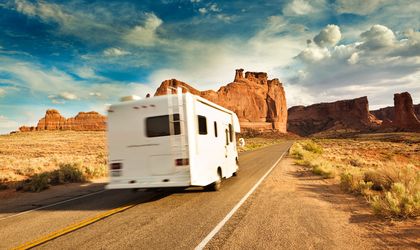 Ein Wohnmobil fährt auf einer Straße durch eine trockene, wüstenähnliche Landschaft mit markanten roten Felsformationen im Hintergrund – Wohnmobil-Abenteuer mit ADAC Reisen.