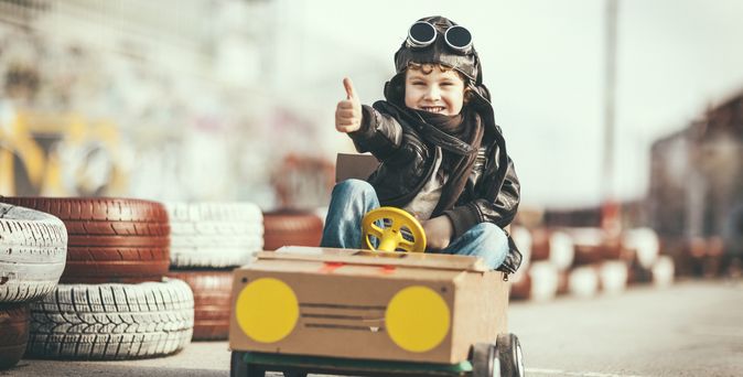 Junge mit Fliegerkappe und Brille gibt Daumen hoch, während er in einem selbstgebastelten Pappauto auf einer angedeuteten Rennstrecke mit Reifenstapeln sitzt.