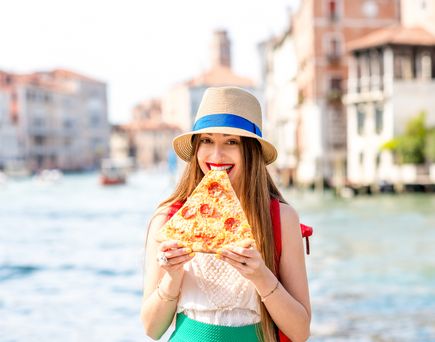 Eine lächelnde Frau mit Hut hält ein großes Stück Pizza vor einem Kanal in Venedig.