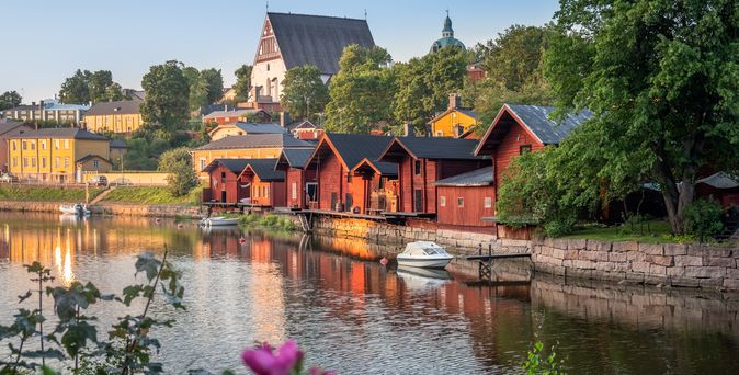 Historische rote Holzhäuser am Flussufer von Porvoo, Finnland, mit einer Kathedrale im Hintergrund und grünen Bäumen unter einem klaren Himmel.