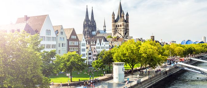 Blick auf die Kölner Altstadt mit Groß St. Martin und dem Kölner Dom im Hintergrund, flankiert vom Rhein und begrünten Uferpromenaden.