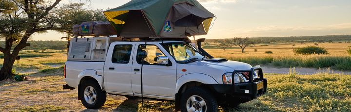 Ein Nissan Navara Pickup-Truck mit einem auf dem Dach montierten Zelt, der auf einer unbefestigten Straße in einer weiten, sonnigen Landschaft steht.