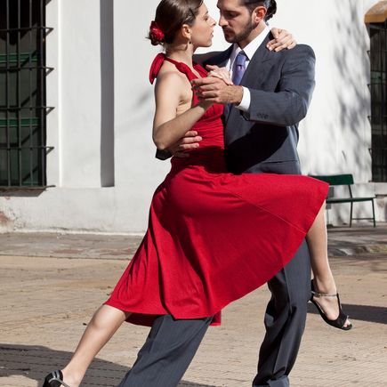 Ein Tanzpaar führt leidenschaftlich Tango in einem Freien auf, mit einer Stadt im Hintergrund und einem Fluss mit Booten.