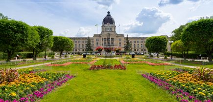 Das Parlamentsgebäude von Saskatchewan in der Hauptstadt Regina. Davor eine weitläufige Wiese, viele angelegte farbenfroh blühenden Blumen, Bäume. Parkähnlich angelegt. Einige Autos parken vor dem Gebäude. Das alles bei blauem Himmel.