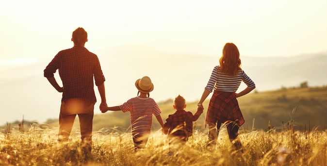 Familie mit Mann, Frau und zwei Kindern halten sich an den Händen und stehen auf einem Feld in herbstlichem Licht. 