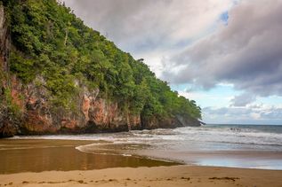 Playa El Valle: Strandansicht mit Wellen, die an den sandigen Uferbereich treffen, links ragt eine steile, mit grüner Vegetation bedeckte Felsklippe auf, unter einem bewölkten Himmel.