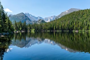 Ein klarer Bergsee spiegelt die umliegenden Nadelwälder und die schroffen Gipfel der Rocky Mountains wider unter einem blauen Himmel mit wenigen Wolken. 