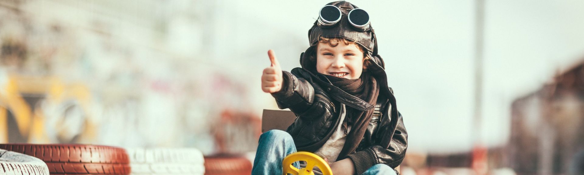 Junge mit Fliegerbrille fährt lachend in einem selbstgebauten Pappkarton-Auto über eine Rennstrecke.