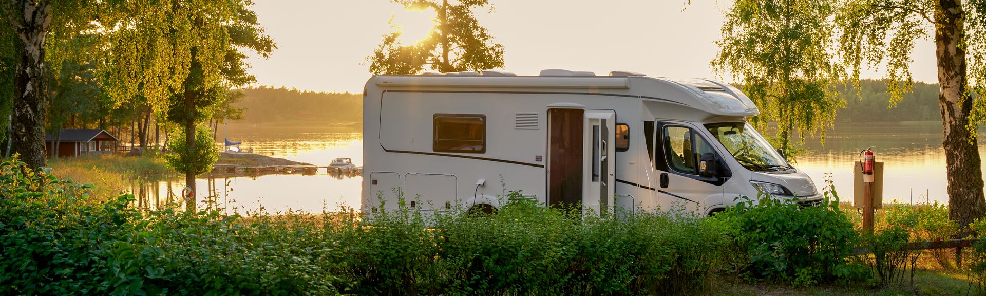 Ein Wohnmobil steht am Ufer eines Sees bei Sonnenuntergang, umgeben von Bäumen und Sträuchern. Im Hintergrund spiegelt sich die untergehende Sonne im Wasser.