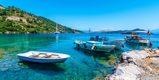 Kleine Boote und ein Segelboot ankern im kristallklaren, türkisfarbenen Wasser der Sarsala Koyu Bucht in Dalaman, Türkei, umgeben von grünen Hügeln.