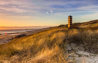 Ein Sandstrand mit Dünen und einem Leuchtturm bei Sonnenuntergang.