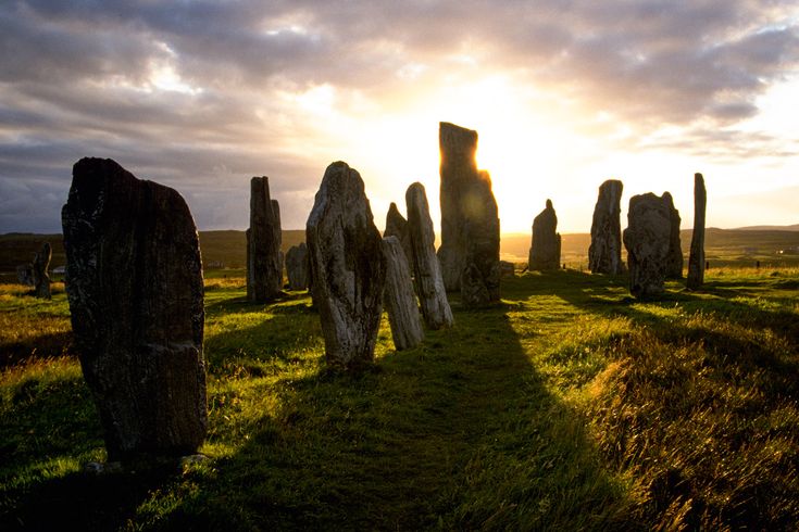 Ein prähistorischer Steinkreis oder eine Reihe von aufrechten Steinen in einer weiten, grasbewachsenen Landschaft bei Sonnenaufgang. Die Sonne scheint durch die Steine und wirft lange Schatten.