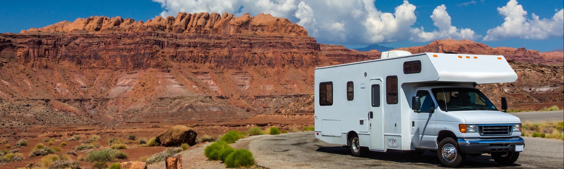 Ein weißes Wohnmobil der Klasse C parkt auf einer asphaltierten Straße inmitten einer trockenen, felsigen Landschaft mit roten Felsformationen im Hintergrund und einem strahlend blauen Himmel mit weißen Wolken