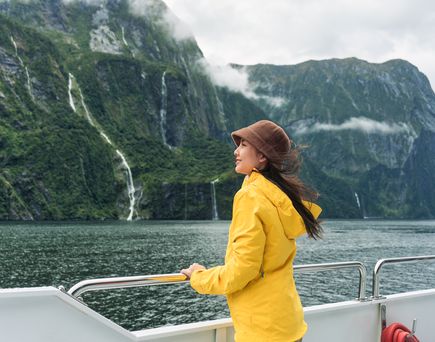Asiatische Frau mit gelben Regenmantel und Hut genießt die Aussicht auf den Milford Sound während einer Fährfahrt im Urlaub in Neuseeland.
