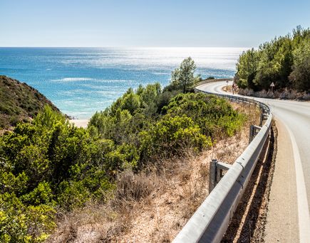 Eine malerische Küstenstraße, die Estrada da Arrábida, schlängelt sich entlang der Küste der Algarve in Portugal, umgeben von grüner Vegetation und dem blauen Atlantik unter einem klaren blauen Himmel.
