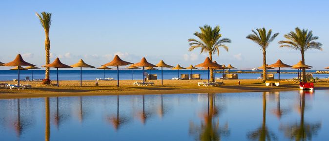 Ruhiger Strand mit Palmen und Strohdächern spiegelt sich idyllisch im glatten Wasser bei strahlend blauem Himmel.