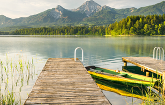 Ein Holzsteg führt zu einem gelb-grünen Ruderboot auf einem ruhigen See in Österreich, umgeben von Bergen.