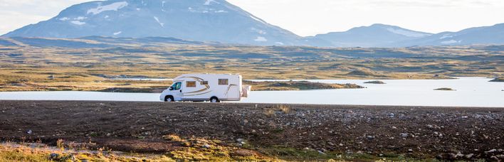 Weißer Campervan fährt auf einem Dammweg, mit Wasser auf der linken und Gras auf der rechten Seite, Berge im Hintergrund.