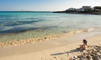 Ein Sandstrand mit Felsen und Meer, ein Baby sitzt im Sand mit blauem Kopftuch.