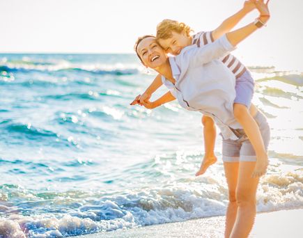 Eine Mutter hat ihren Sohn auf dem Rücken, am Strand, das Wasser ist zu sehen und die große Freude, welche beide verspüren.